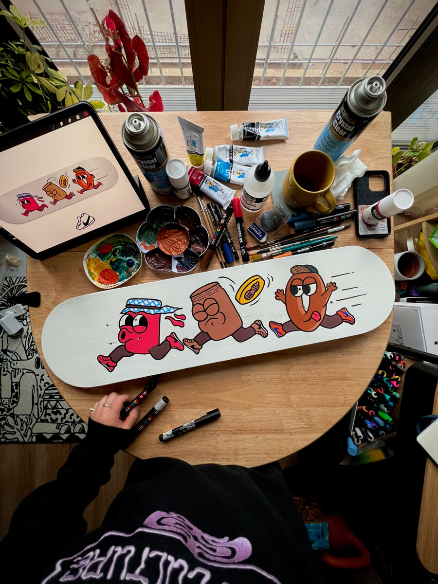 peanut butter character chased by jam jar and bagel characters painted on a white skate deck